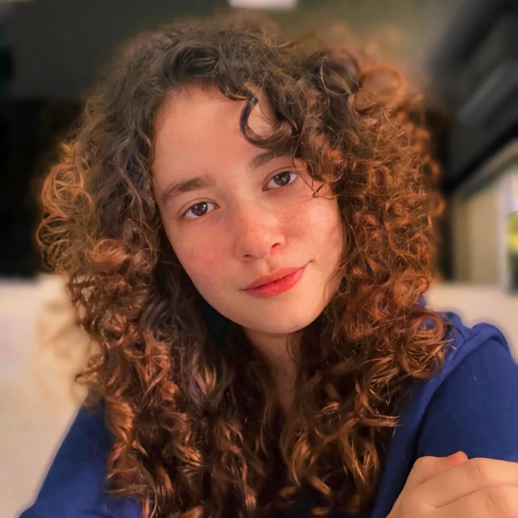 Curly-haired dark eyes woman in blue shirt, smiling gently, with hand resting on arm.